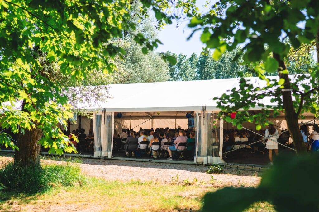 Een bijzondere vergaderlocatie in de natuur met een aluhal waar mensen in moderne sfeer luisteren naar een spreker – even wat anders als een strand locatie, rolstoeltoegankelijk en perfect voor een congrescentrum met capaciteit op maat.